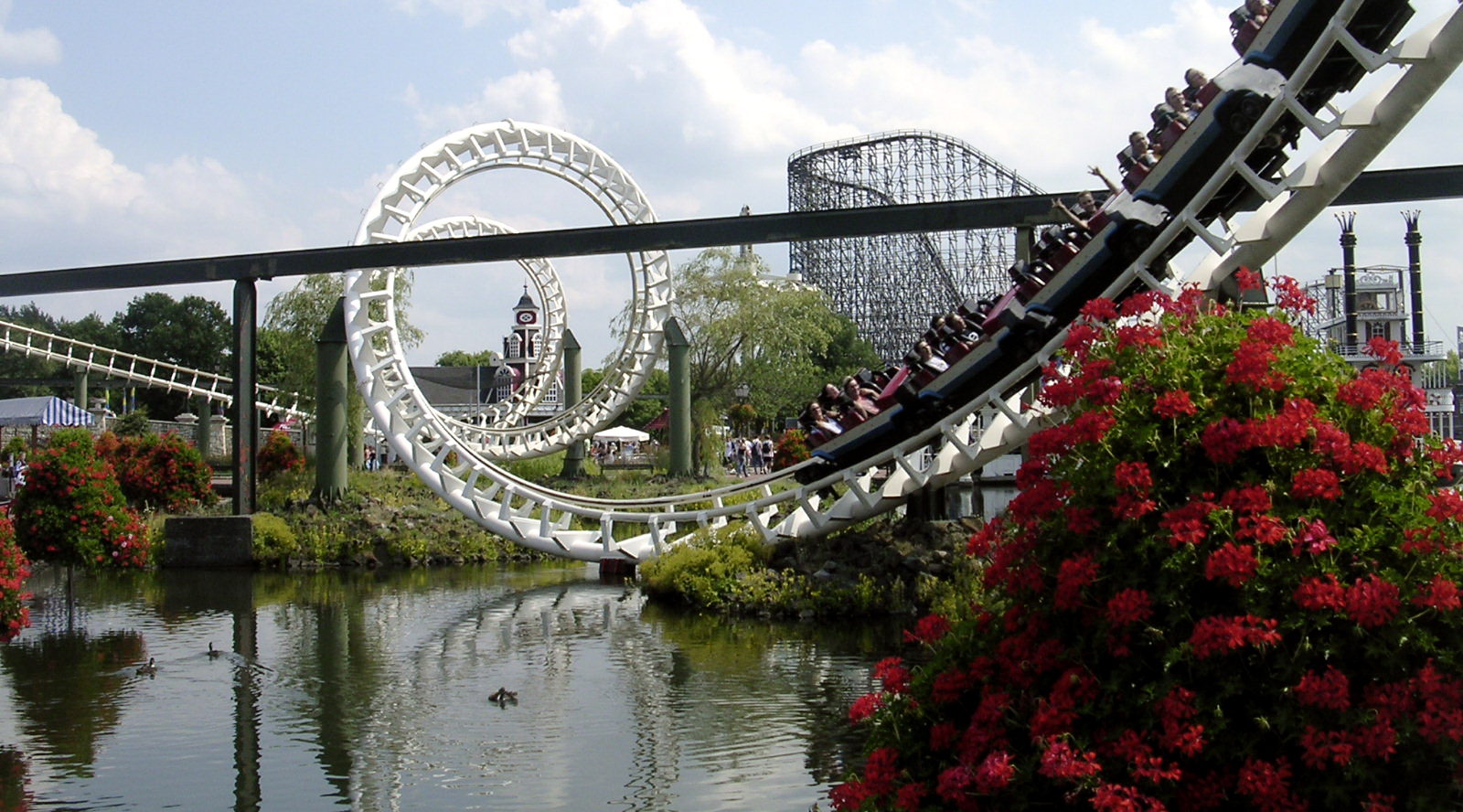 A white roller coaster in an amusement park with red flowers in the background.